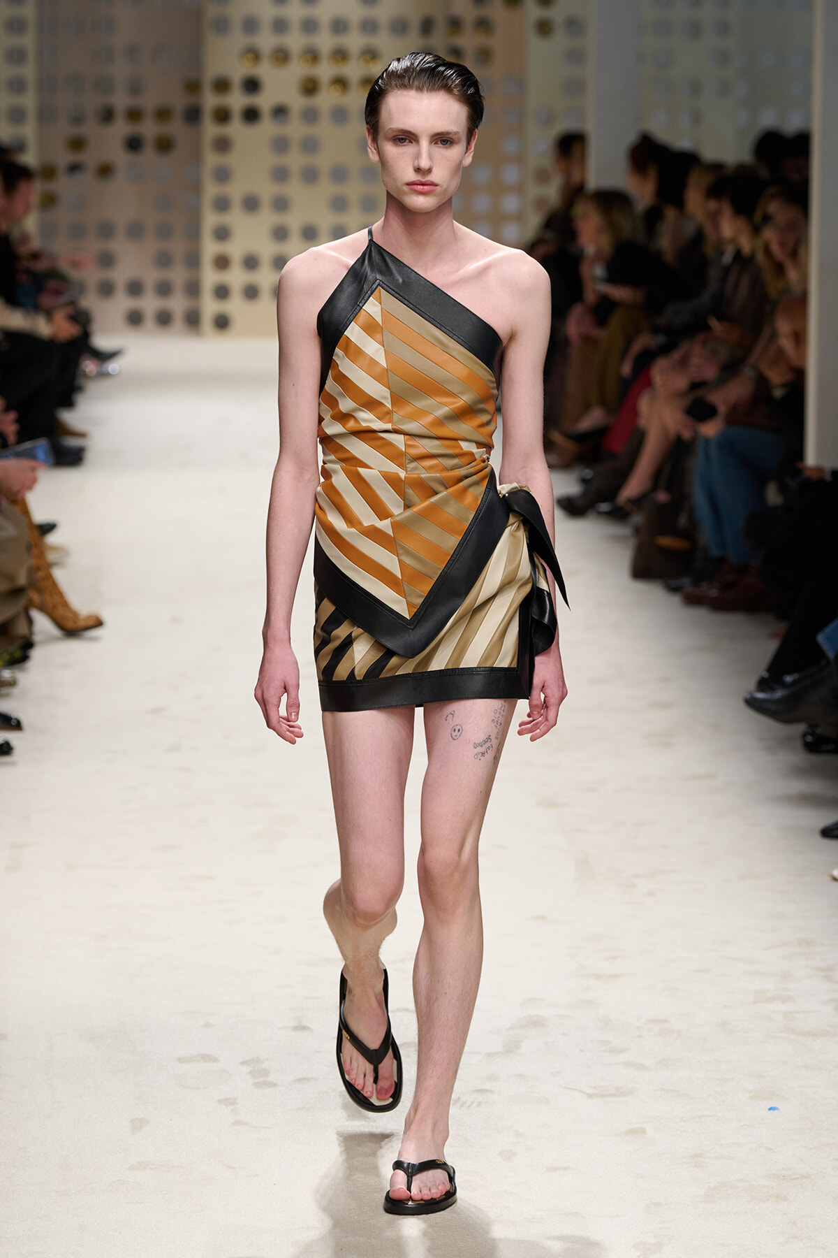 Female runway model wearing a tan, gold, and black geometric one-shoulder mini dress with black trim, walking the catwalk in sandals.