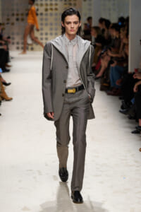 Model walking a fashion runway in a gray suit with a pale shirt and light-gray hooded layer, audience seated along the sides.