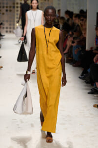 Female model walks a fashion runway in a mustard yellow sleeveless jumpsuit, carrying a white handbag and wearing brown sandals, with an audience in the background.