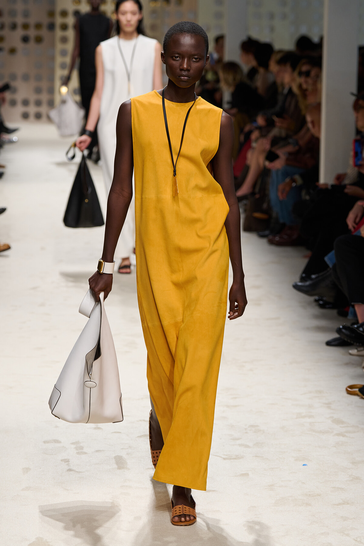 Female model walks a fashion runway in a mustard yellow sleeveless jumpsuit, carrying a white handbag and wearing brown sandals, with an audience in the background.