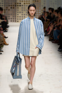 Model walks a fashion runway in a blue and white striped oversized shirt over a cream shift dress, carrying a matching striped tote bag.