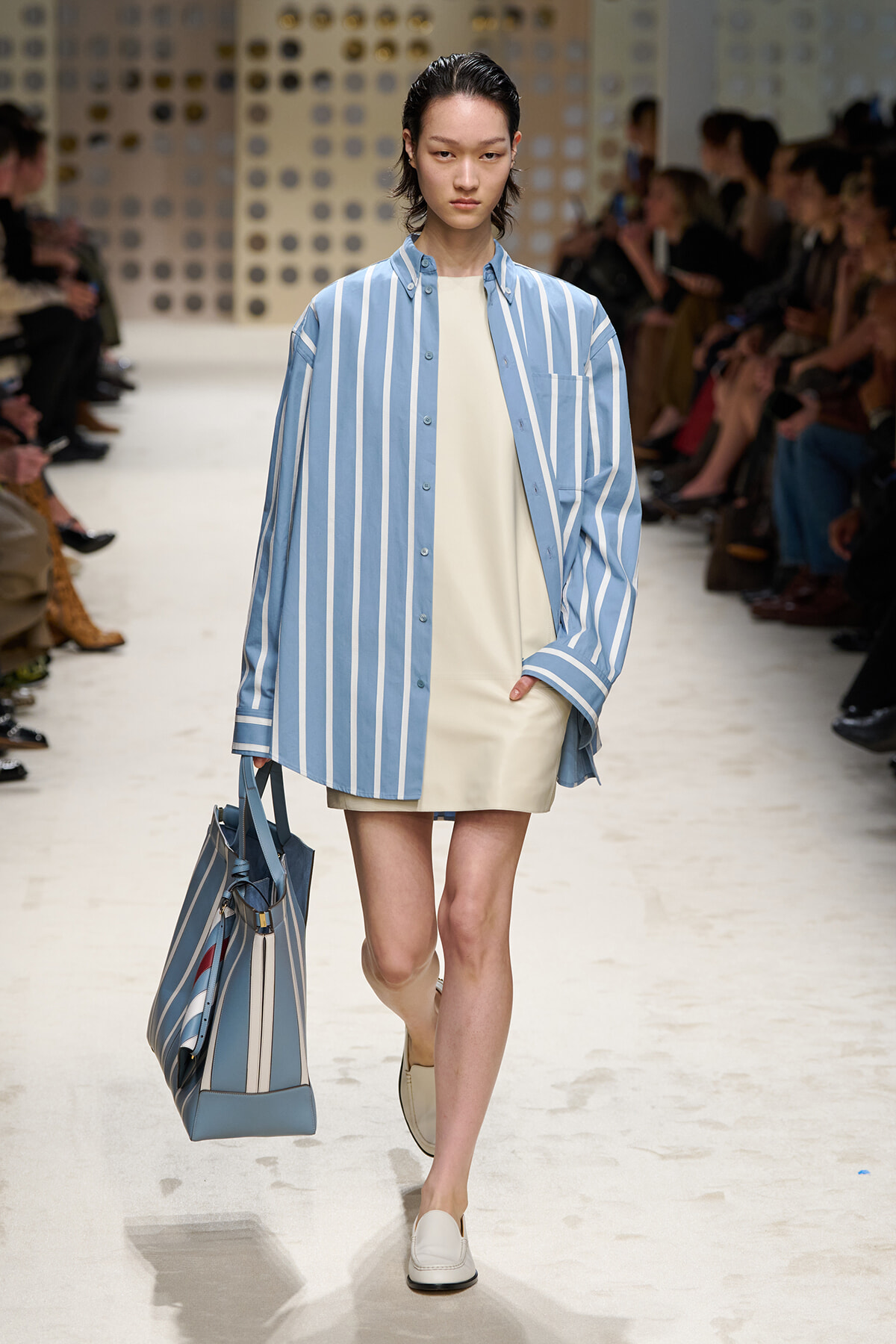 Model walks a fashion runway in a blue and white striped oversized shirt over a cream shift dress, carrying a matching striped tote bag.