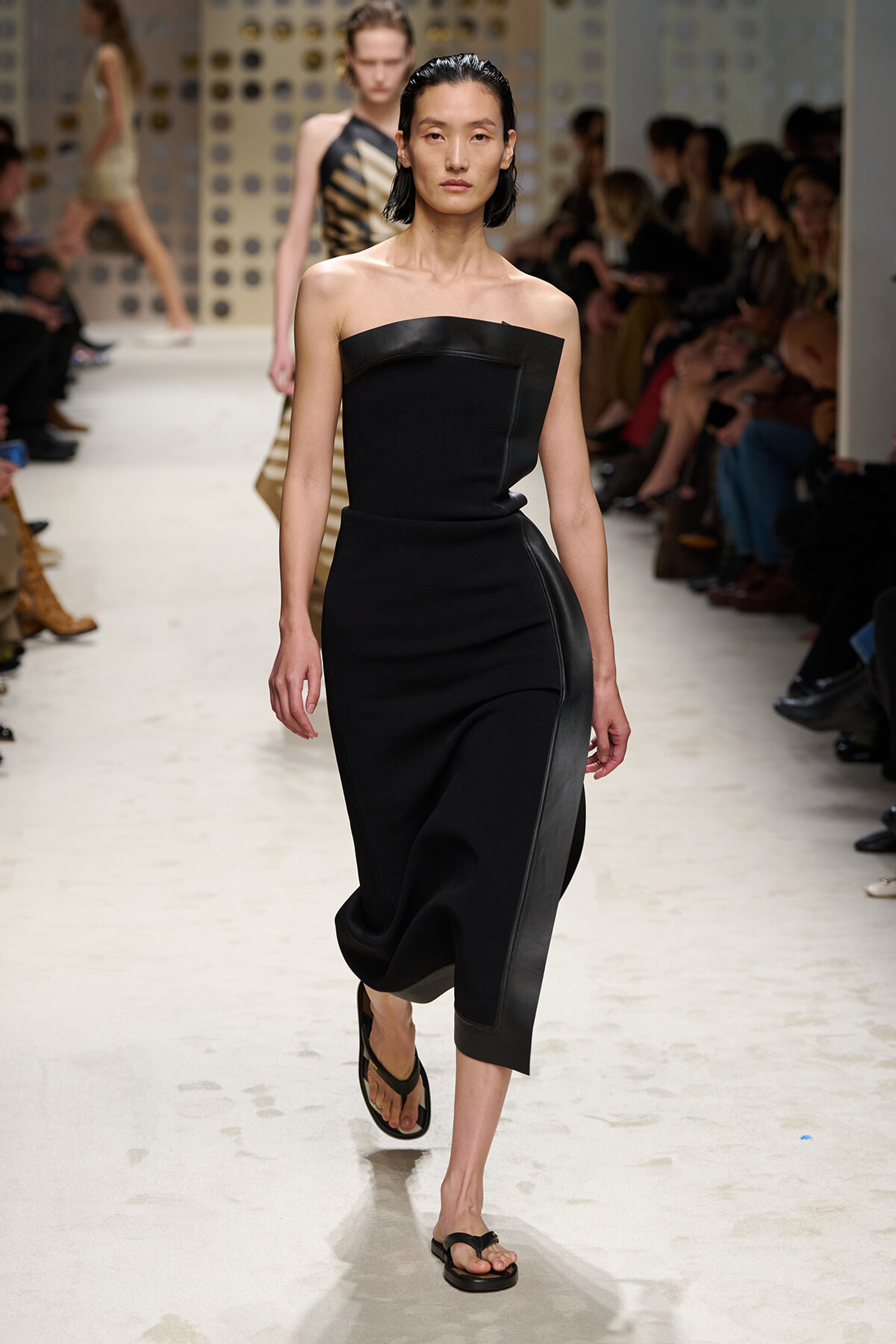 Female model walks the runway in a strapless black evening dress with asymmetrical neckline and sleek belt-like seam, wearing flip-flop sandals.