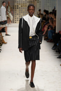 Female model walks a runway in a black pinstripe suit with a white shirt and oversized white-collar jacket, audience in background