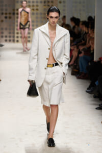 Model walks the runway in a white oversized blazer and matching knee-length shorts, unbuttoned at the chest, with a black belt and black loafers, carrying a small black handbag. Audience visible in the background.