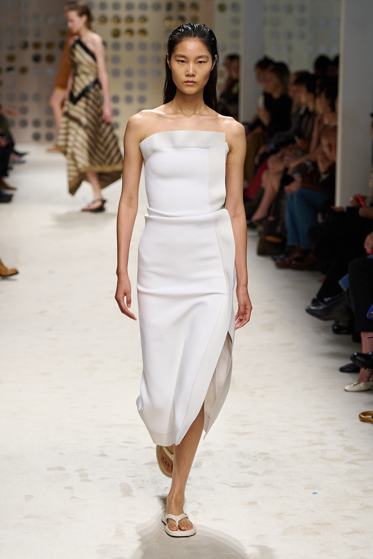 Model walks the runway in a white strapless midi dress with a front panel and slit.