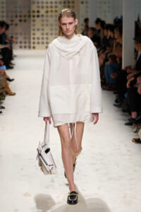Model walks the runway in an oversized white hooded dress with large front pocket, carrying a white handbag; black embellished loafers complete the look.