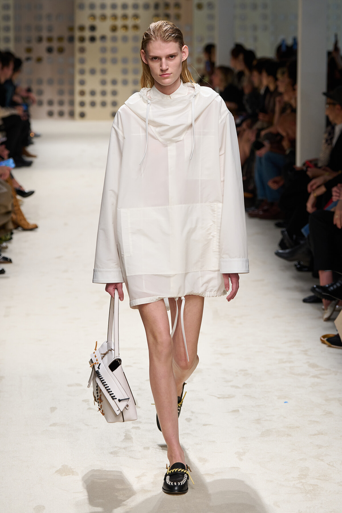 Model walks the runway in an oversized white hooded dress with large front pocket, carrying a white handbag; black embellished loafers complete the look.