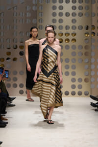 Model wearing a beige and black striped one-shoulder dress walks the runway with a perforated backdrop behind her.