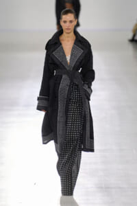 Model walking a runway in a black-and-white patterned trench coat with a belt over polka-dot pants.