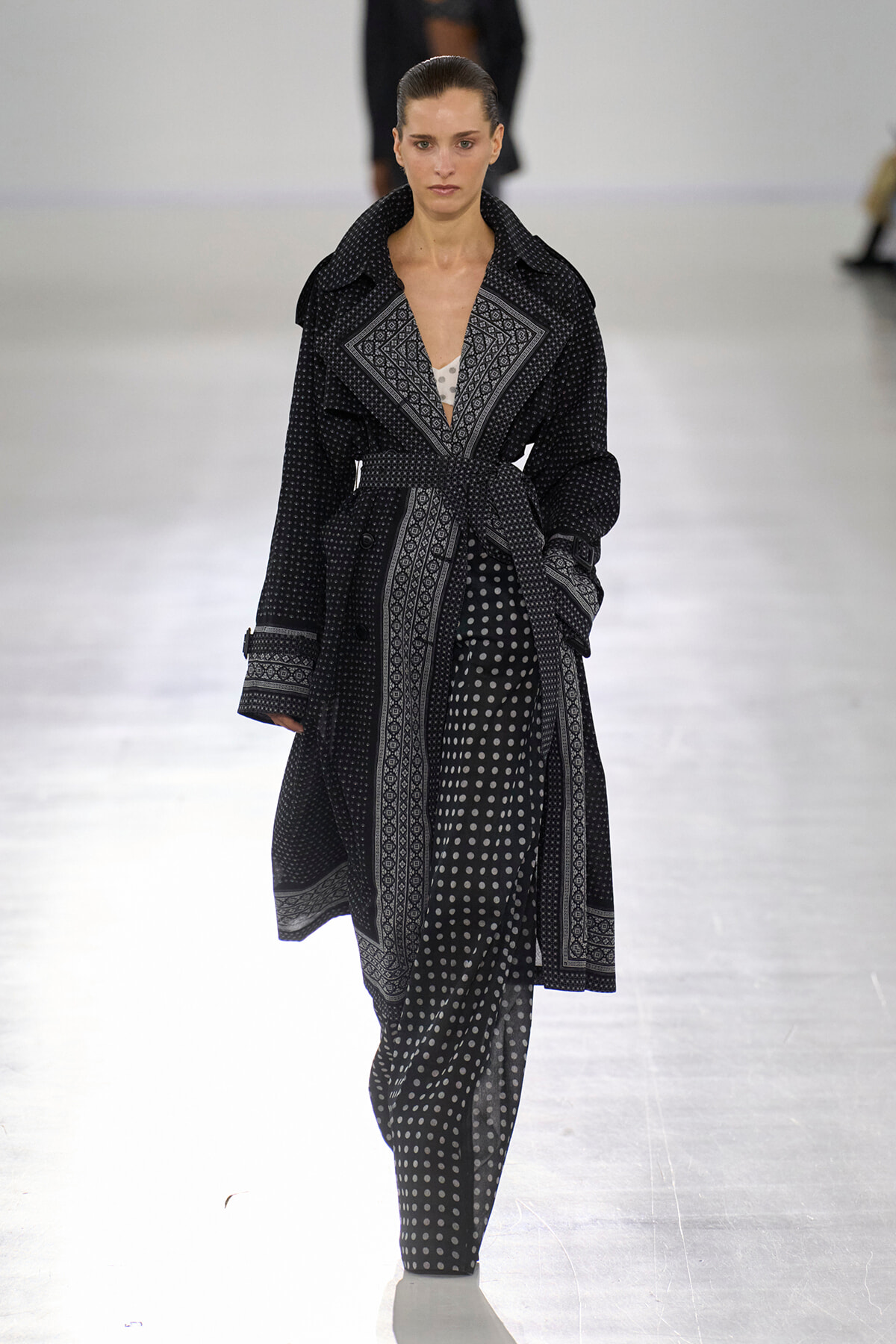 Model walking a runway in a black-and-white patterned trench coat with a belt over polka-dot pants.