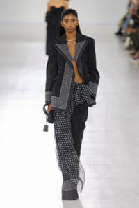 Female model walks the runway in a black-and-white patterned suit with a geometric blazer and matching wide-leg pants, holding a small black handbag.