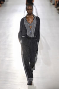 Model walking a fashion runway in a black-and-white patterned jumpsuit with a belt and pearl necklace, hands in pockets.