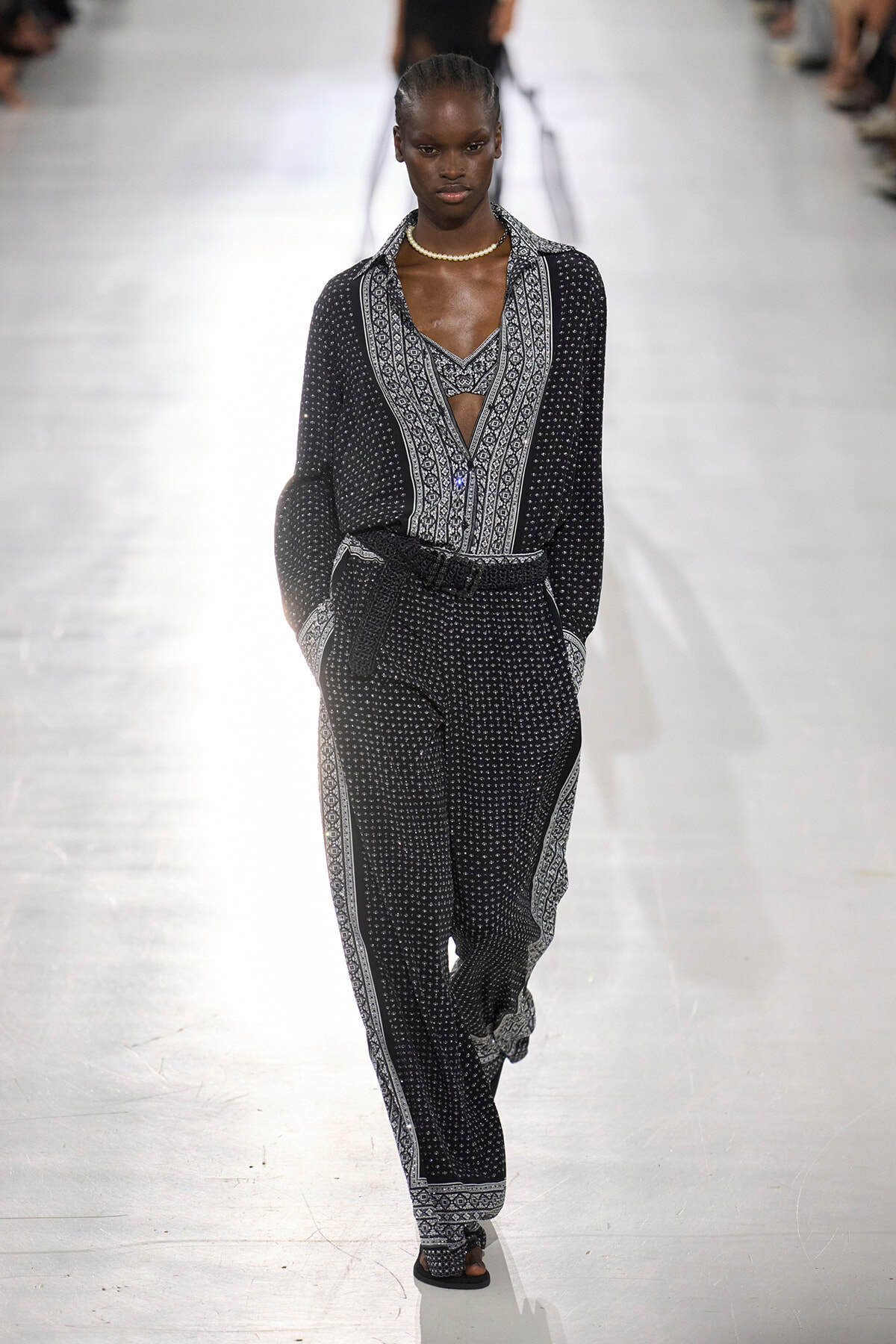Model walking a fashion runway in a black-and-white patterned jumpsuit with a belt and pearl necklace, hands in pockets.
