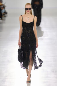 Model on a runway wearing a black lace slip dress with thin straps, oversized sunglasses, and a pearl necklace, carrying a black handbag.