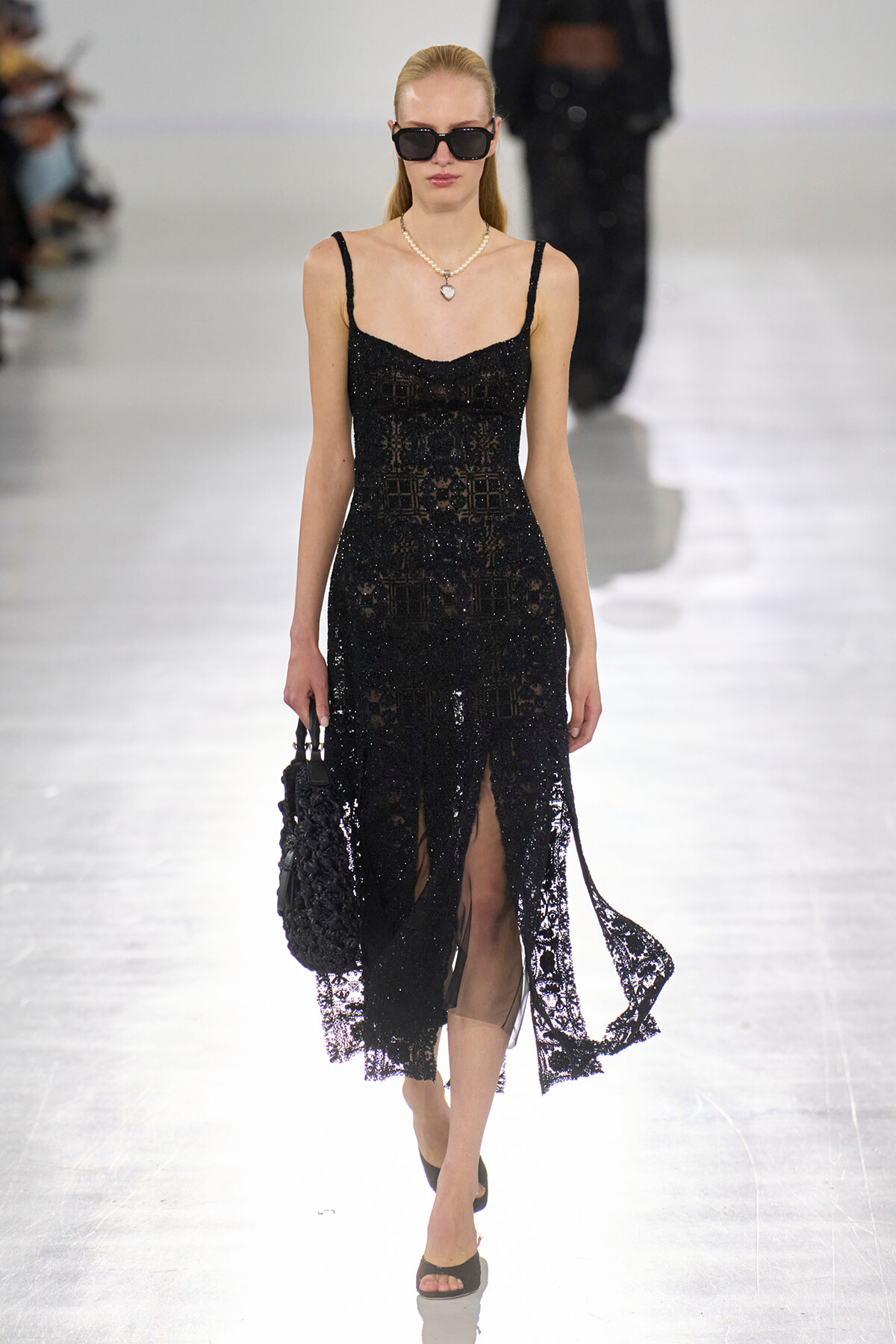 Model on a runway wearing a black lace slip dress with thin straps, oversized sunglasses, and a pearl necklace, carrying a black handbag.