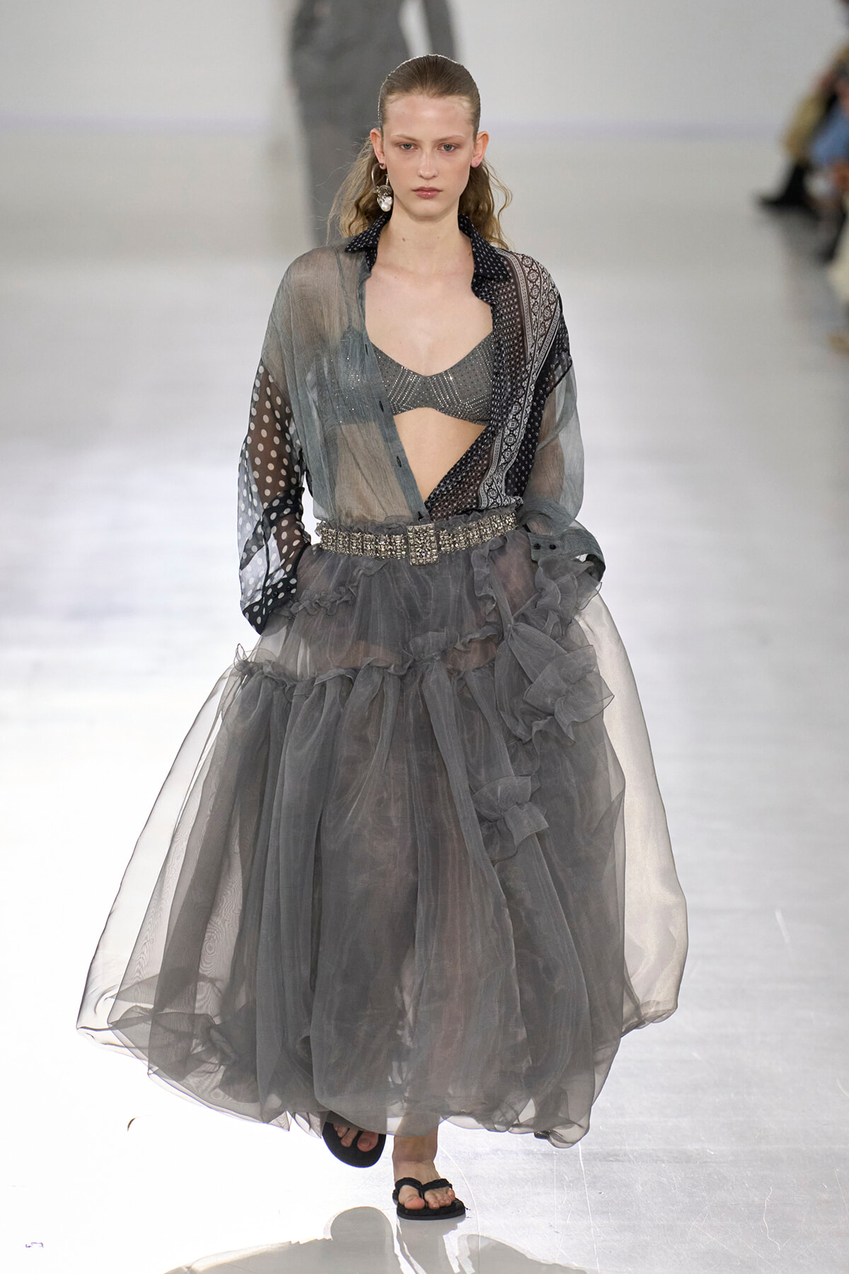 Runway model in a sheer gray blouse over a metallic bralette, with a voluminous layered tulle skirt and jeweled belt.