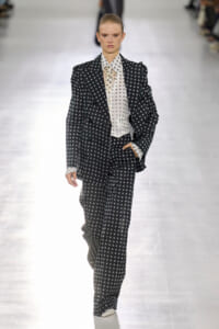 Fashion model walks the runway in a black polka-dot suit with a white polka-dot shirt and matching trousers.