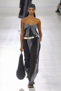 Runway model wearing a black-and-white printed strapless top and wide-leg pants with a white belt, polka-dot headscarf, and a large black textured bag.
