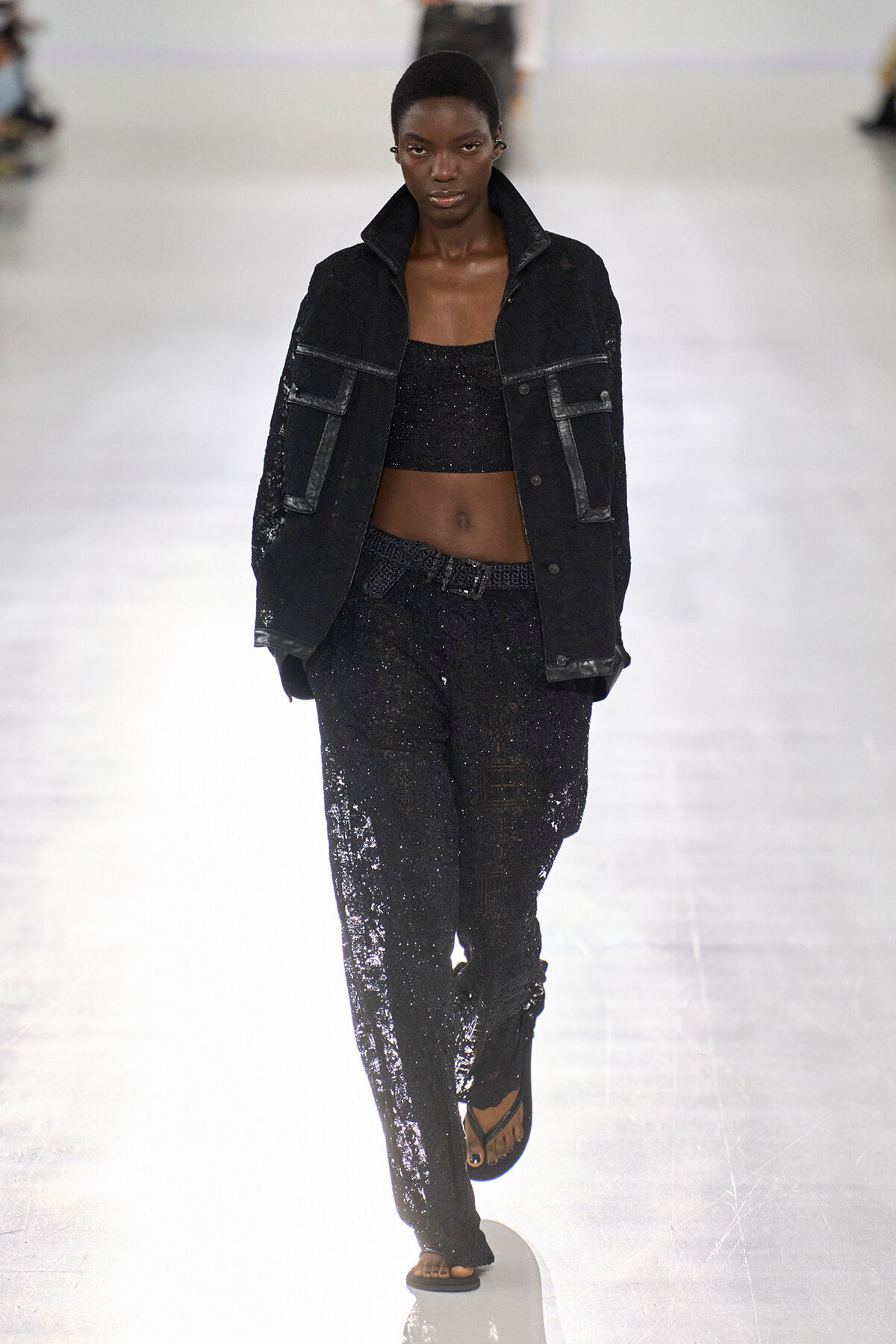 Model walks the runway in a black sequined crop top and matching wide-leg sequined pants, paired with an oversized black denim jacket with large pockets.
