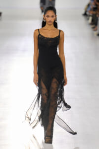 Model walking the runway in a black lace slip dress with sheer overlay and thin straps, wearing statement earrings.
