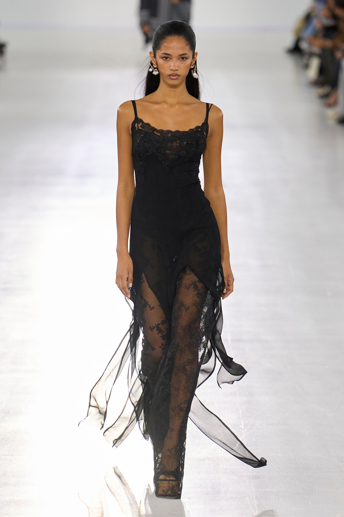 Model walking the runway in a black lace slip dress with sheer overlay and thin straps, wearing statement earrings.