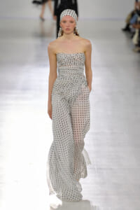 Model walking a fashion runway in a strapless, ruched, white and gray polka-dot jumpsuit with a matching headscarf and dangling earrings.