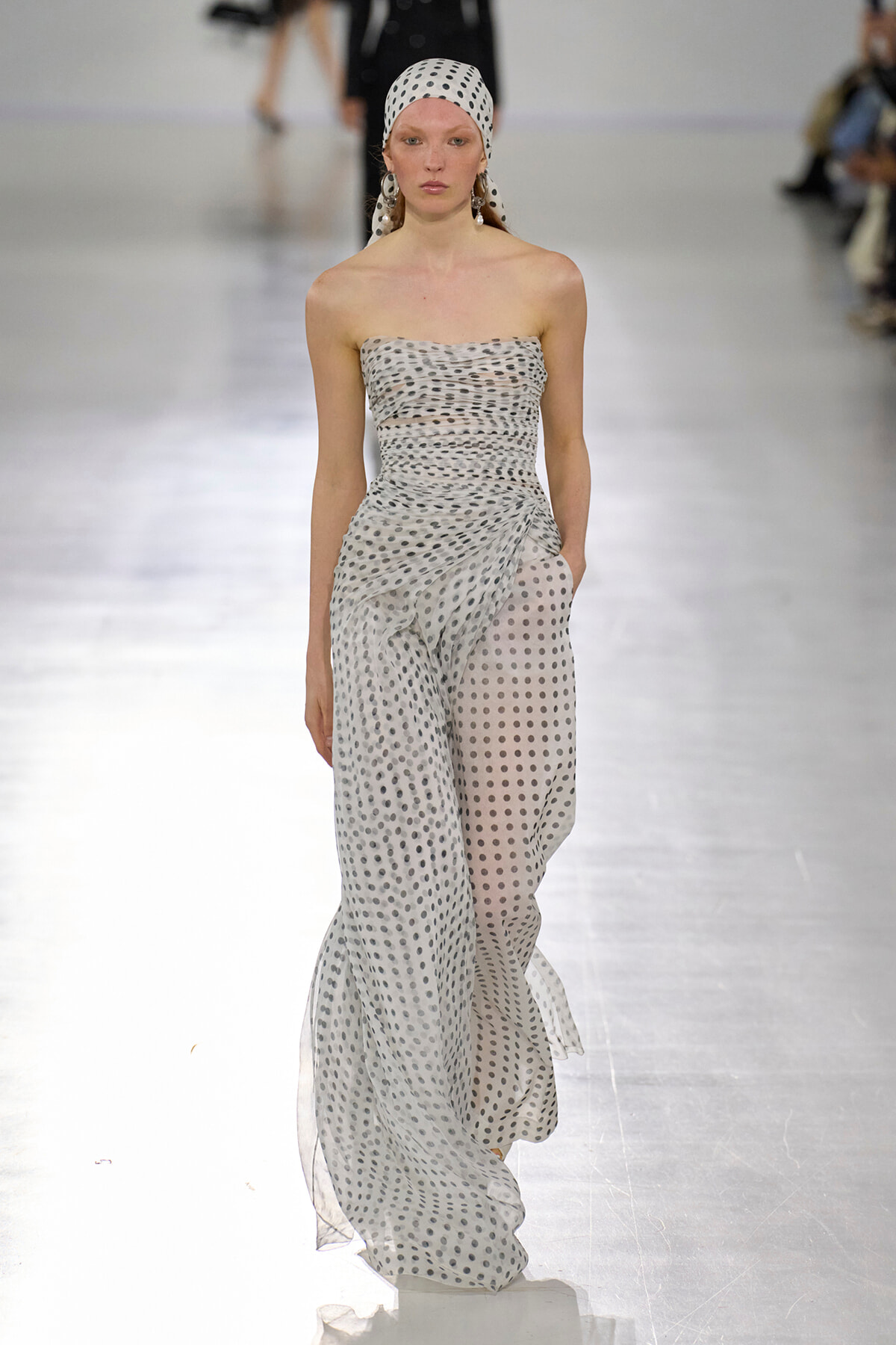 Model walking a fashion runway in a strapless, ruched, white and gray polka-dot jumpsuit with a matching headscarf and dangling earrings.