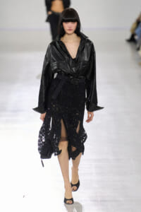 Model walking a runway in a black leather blouse tucked into a glittery black midi skirt with a belt and open-toed heels.