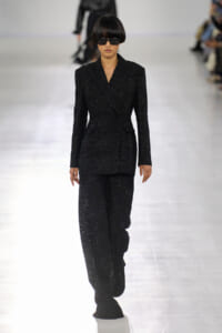 Female model walks the runway in a black sparkling pantsuit with a matching blazer and wide sunglasses, on a bright runway.