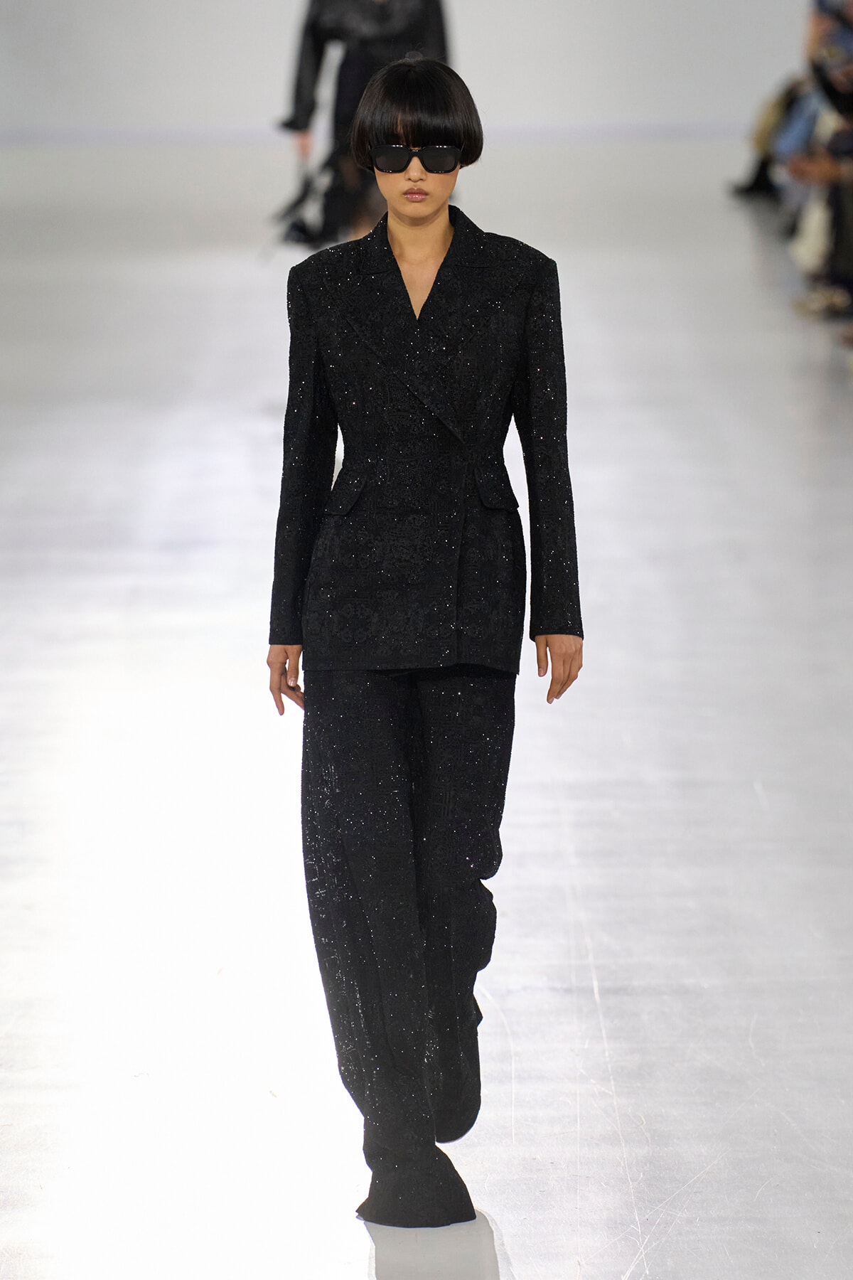 Female model walks the runway in a black sparkling pantsuit with a matching blazer and wide sunglasses, on a bright runway.