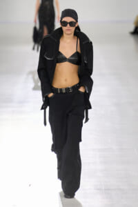 Model on runway wearing a black bralette, oversized jacket, and cargo pants with a belt and sunglasses, hands in pockets.