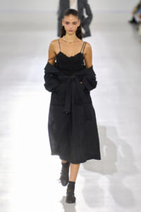 Model walking a fashion runway in a black lace spaghetti-strap dress with a draped black coat tied at the waist and a pearl necklace.