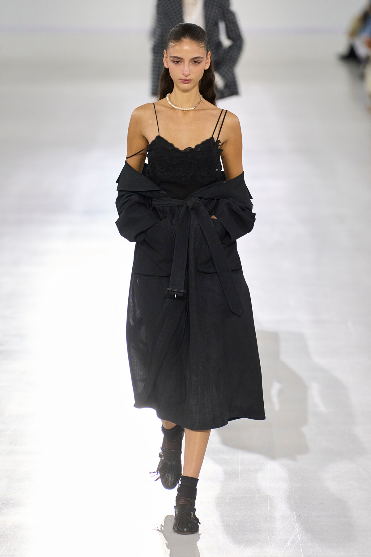 Model walking a fashion runway in a black lace spaghetti-strap dress with a draped black coat tied at the waist and a pearl necklace.