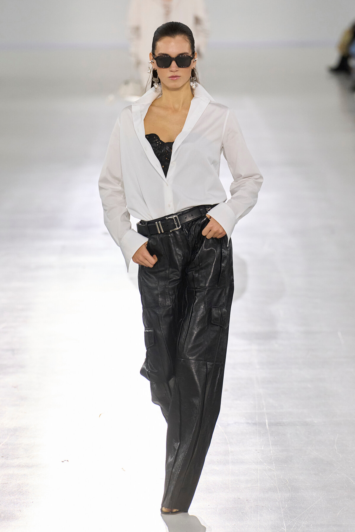 Model walking a runway in a white button-down shirt, black lace top, and loose black cargo pants with a belt, wearing dark sunglasses.
