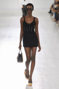 Runway model wearing a black knit mini dress with decorative panels, holding a small black handbag with studded edges and wearing sunglasses.
