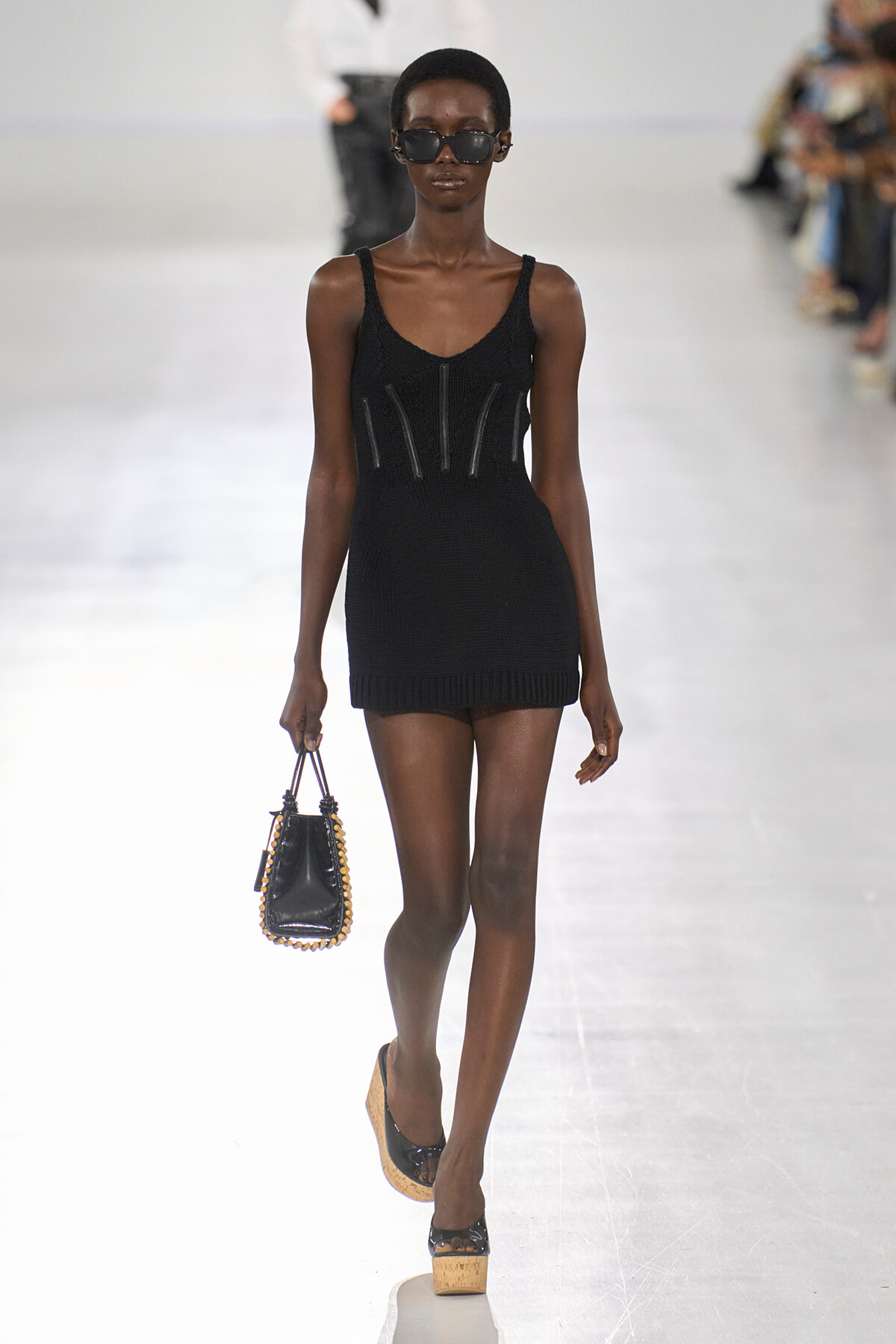 Runway model wearing a black knit mini dress with decorative panels, holding a small black handbag with studded edges and wearing sunglasses.
