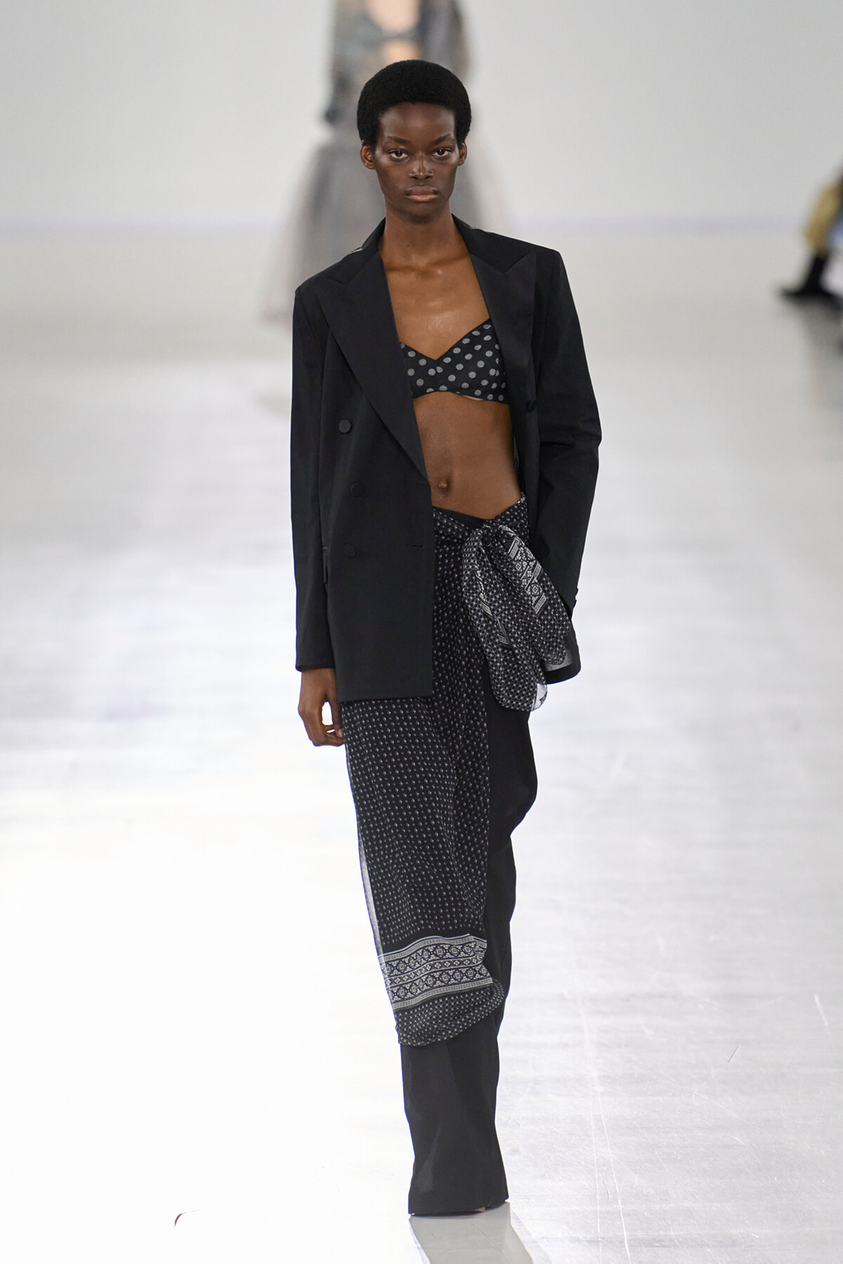 Model walking a runway in a black blazer over a polka-dot bra, with black patterned scarf belt and wide-leg pants.
