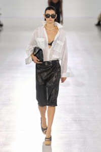 Model on a runway wearing a sheer white blouse over a black lingerie top, a black leather midi skirt, belt, pearls, and oversized sunglasses, carrying a black clutch.