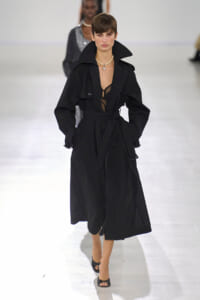 Model on a runway wearing a long black trench coat with a cinched waist and wide collar, paired with a pearl necklace and open-toe heels.