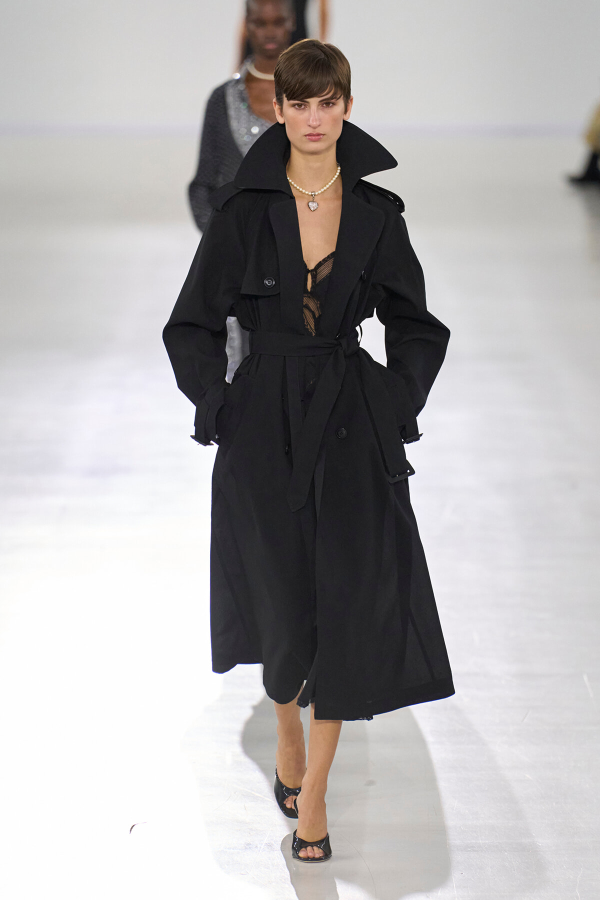 Model on a runway wearing a long black trench coat with a cinched waist and wide collar, paired with a pearl necklace and open-toe heels.