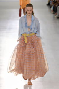 Model walks a runway in a light blue patterned blouse tucked into a voluminous orange plaid skirt with a yellow belt. Audience blurred in background.