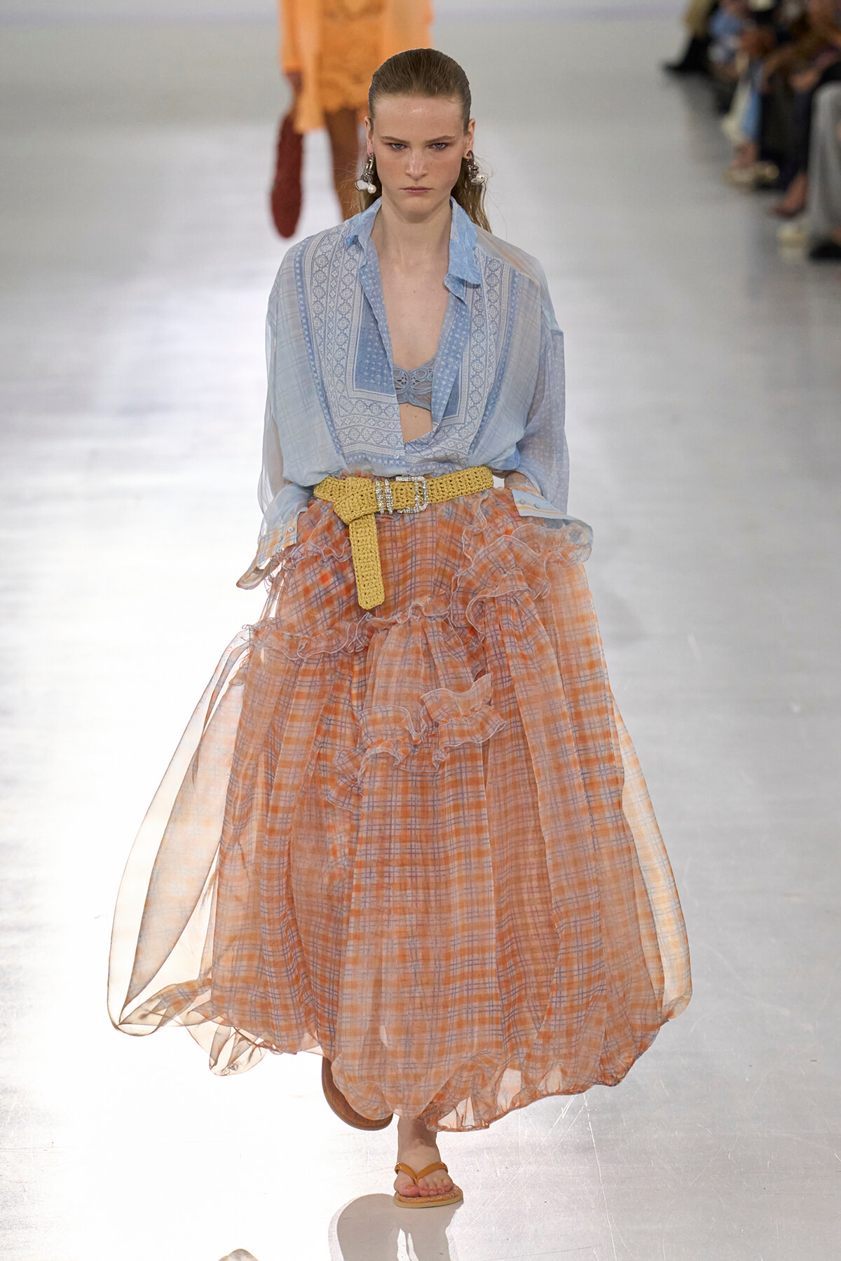Model walks a runway in a light blue patterned blouse tucked into a voluminous orange plaid skirt with a yellow belt. Audience blurred in background.