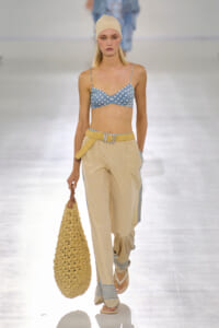 Runway model wearing a blue polka-dot bralette with beige patterned wide-leg pants, a yellow knit belt, a matching knit cap, and a large crocheted tote.