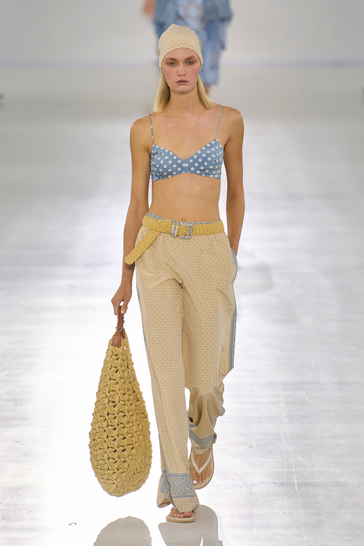Runway model wearing a blue polka-dot bralette with beige patterned wide-leg pants, a yellow knit belt, a matching knit cap, and a large crocheted tote.
