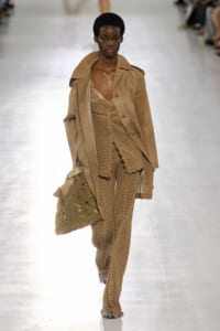 Runway model in a beige crochet-knit ensemble with a matching trench coat, carrying a large crocheted bag.