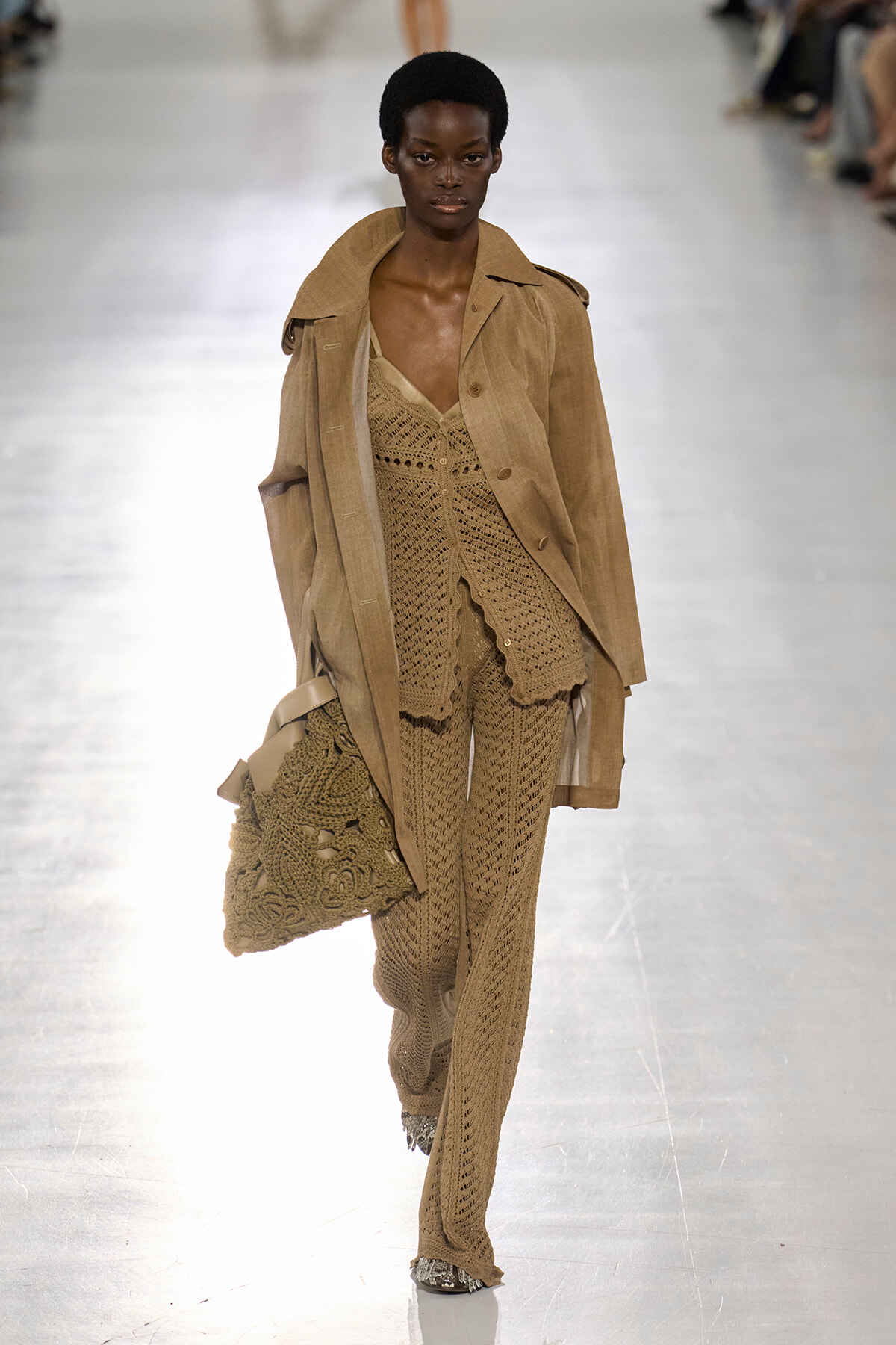 Runway model in a beige crochet-knit ensemble with a matching trench coat, carrying a large crocheted bag.
