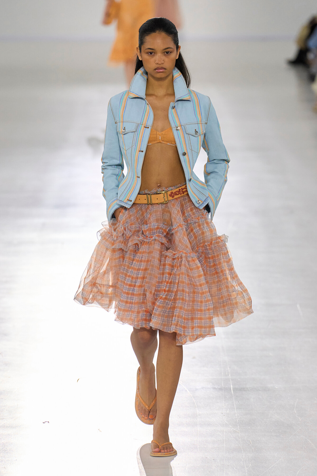 Model walking the runway in a light blue striped denim jacket, orange bralette, and a layered peach plaid tulle skirt with a tan belt.