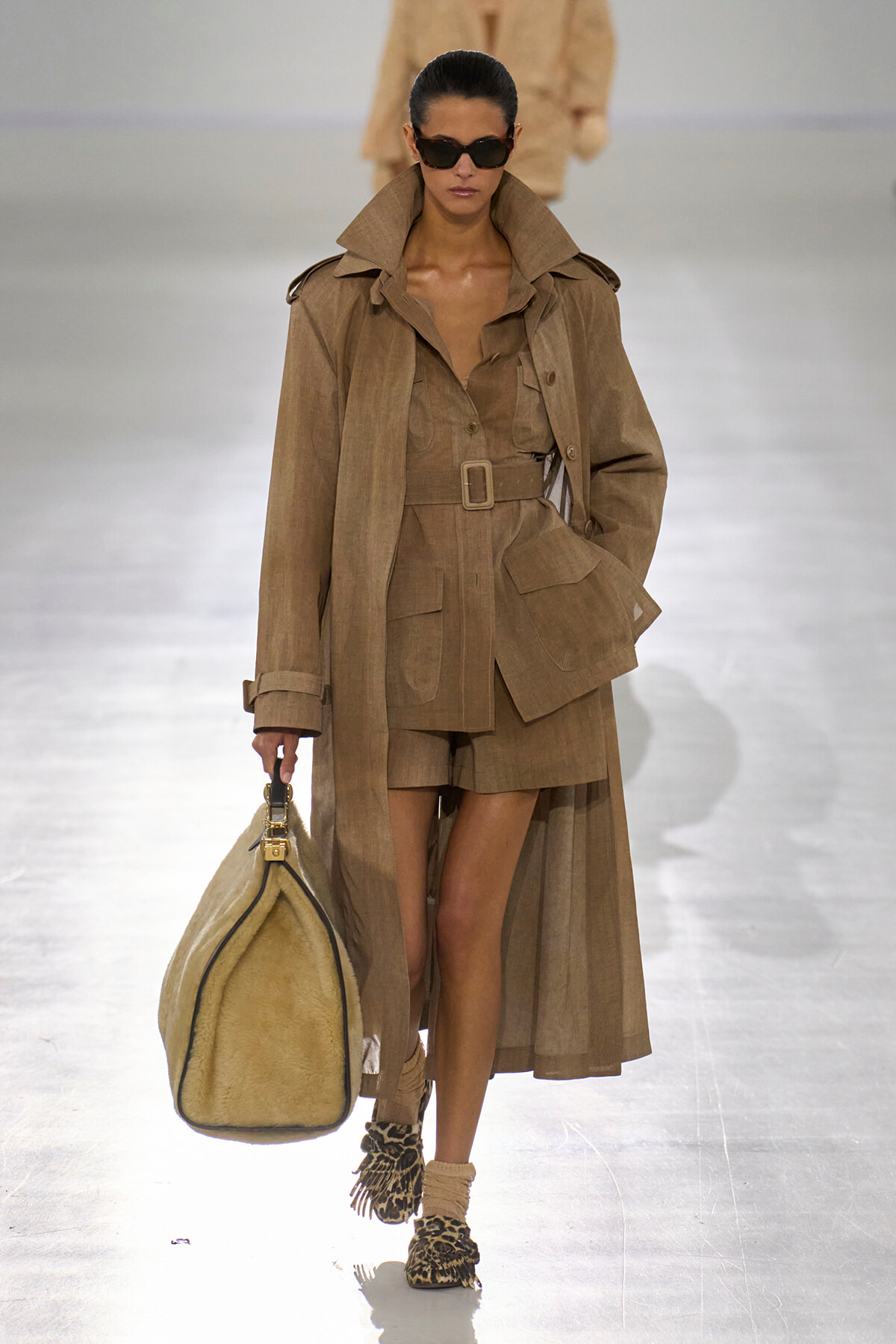 Model walks the runway in a tan belted trench coat with matching shorts and a large beige duffel bag in hand, wearing dark sunglasses.