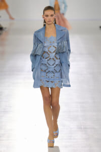 Runway model wearing a light-blue lace mini dress with a matching blue fringe-embellished jacket and platform wedges.